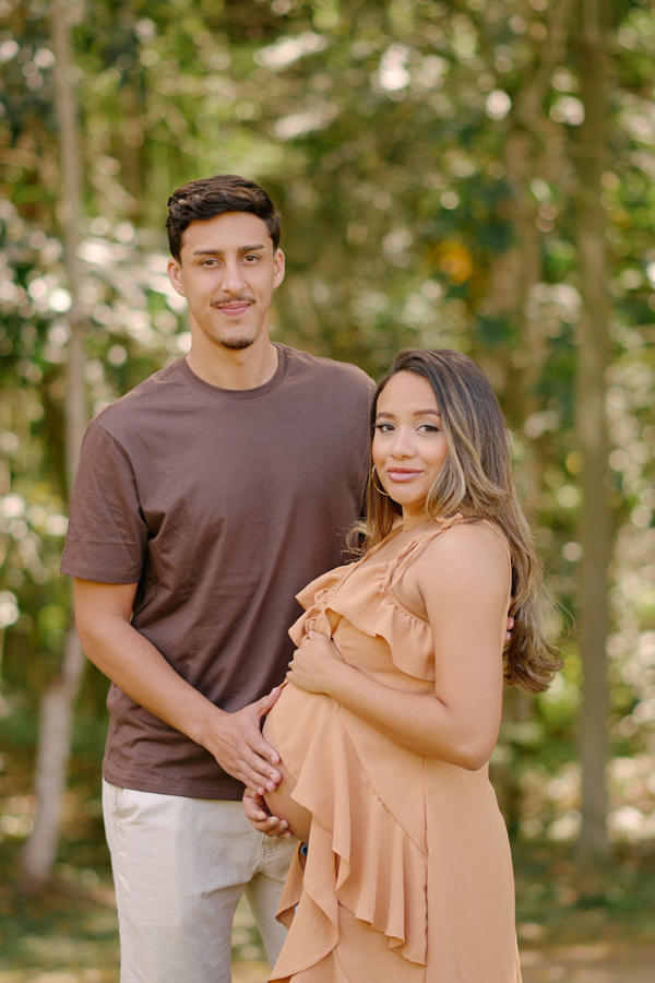 Ensaio gestante em São José dos Campos. Felipe e Lorrana posam sorridentes entre as árvores do Parque da Cidade. Ela usa um vestido pêssego que destaca a barriga à espera de Pedro, em um registro de maternidade com cores quentes e harmonia natural.