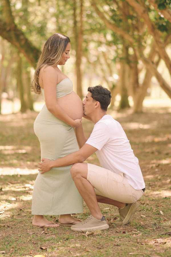 Ensaio gestante em São José dos Campos. Felipe ajoelhado beija a barriga de Lorrana em um momento de carinho à espera de Pedro. O casal usa tons claros e está cercado pelas árvores do Parque da Cidade, transmitindo paz e a doçura da espera.