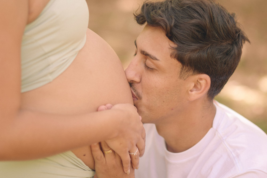Close emocionante do ensaio gestante em São José dos Campos. Felipe beija a barriga de Lorrana com os olhos fechados, demonstrando carinho e a doce espera por Pedro. A luz suave do Parque da Cidade destaca a conexão e o amor desse momento único.
