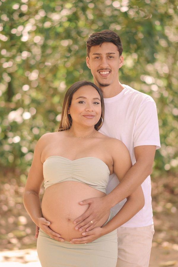 Ensaio gestante em São José dos Campos. Close de Lorrana e Felipe sorridentes, com ele a abraçando por trás e ambos com as mãos na barriga à espera de Pedro. O registro no Parque da Cidade destaca a felicidade e o brilho do casal em tons claros.