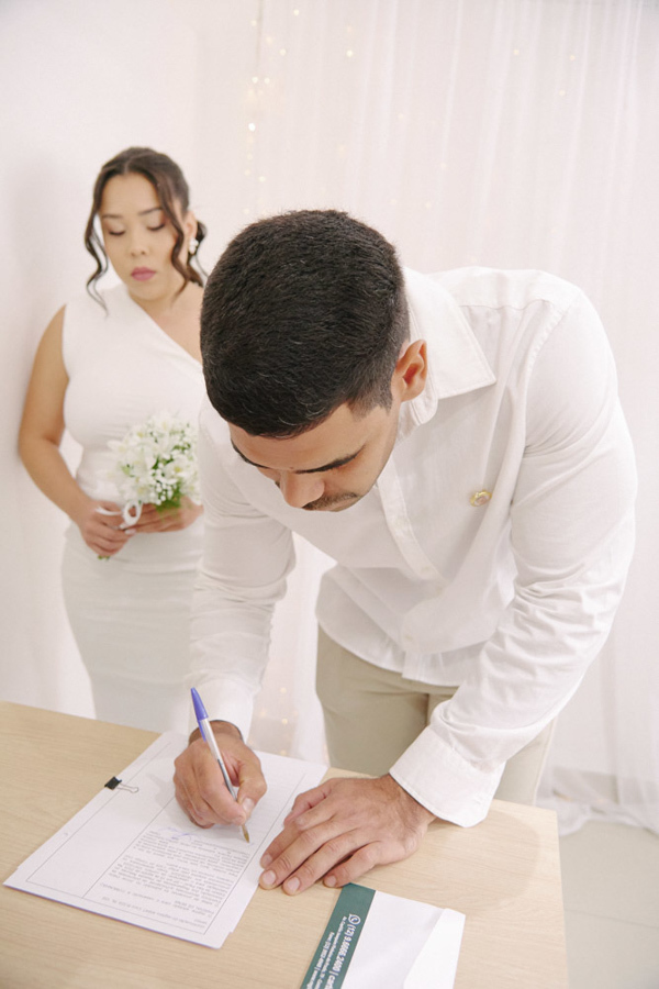 Fotografia de casamento civil em SJC: Luiz Carlos assina a documentação no cartório enquanto Gabrielle observa ao fundo. Registro documental focado na importância do 'sim' oficial e na construção da nova família em São José dos Campos.