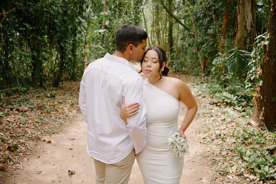 Fotografia de casamento em SJC: Luiz Carlos beija a testa de Gabrielle em uma trilha cercada por vegetação densa. O registro captura um momento de proteção e carinho profundo durante o ensaio pós-civil em São José dos Campos.