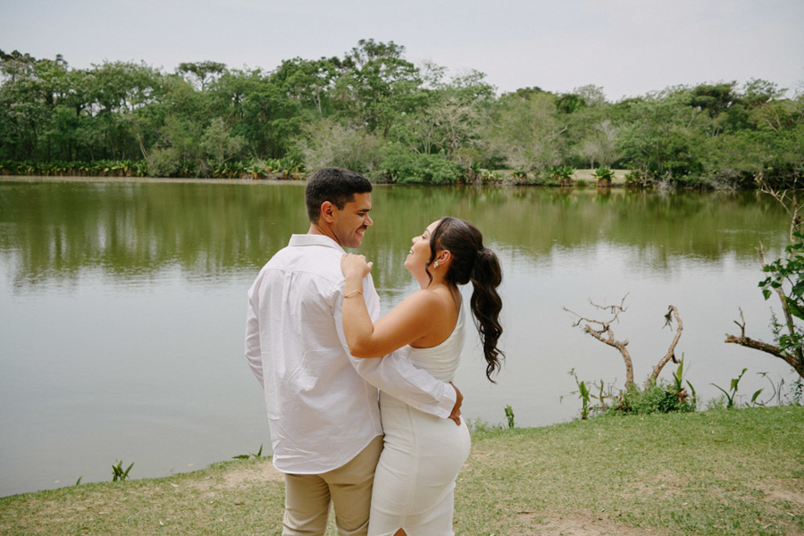 Fotografia de casamento em SJC: Gabrielle e Luiz Carlos sorriem um para o outro em frente ao lago do Parque da Cidade. O ensaio pós-civil em São José dos Campos destaca a felicidade do casal em um cenário natural sereno e apaixonante.