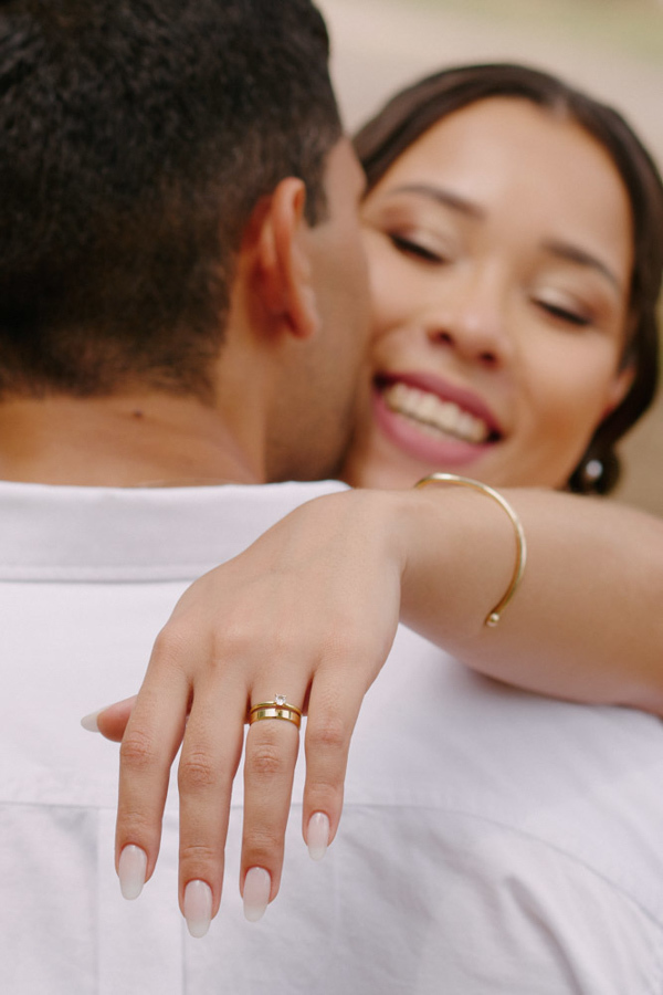 Fotografia de casamento em SJC: Close-up da mão de Marcela com a aliança de ouro sobre o ombro de Fernando. O registro foca no detalhe da joia e na expressão de felicidade da noiva durante o ensaio romântico em São José dos Campos.