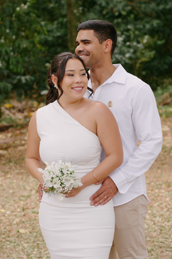 Fotografia de casamento em Jacareí: Fernando abraça Marcela por trás em momento de pura alegria. O registro destaca o sorriso do casal e o buquê branco, capturando a felicidade radiante durante o ensaio pós-civil em um cenário natural.