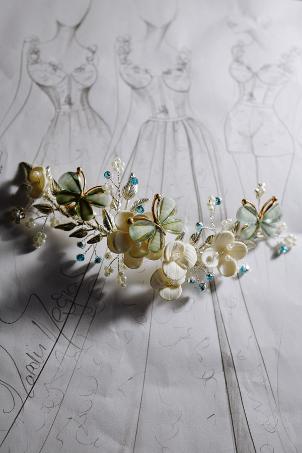 Acessório de cabelo artesanal com borboletas verdes e flores brancas repousa sobre croquis de vestidos desenhados à mão. Detalhe delicado que une a moda ao talento artístico da debutante Anna Clara em Jacareí. Fotografia com luz suave e sombras.