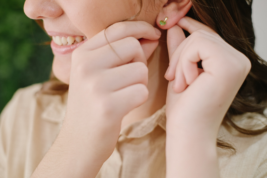 Close documental de Anna Clara sorrindo enquanto coloca um brinco verde. O registro captura a sensibilidade e os preparativos da debutante em Jacareí, focando em gestos espontâneos e na expressão autêntica de quem vive a arte em cada detalhe.