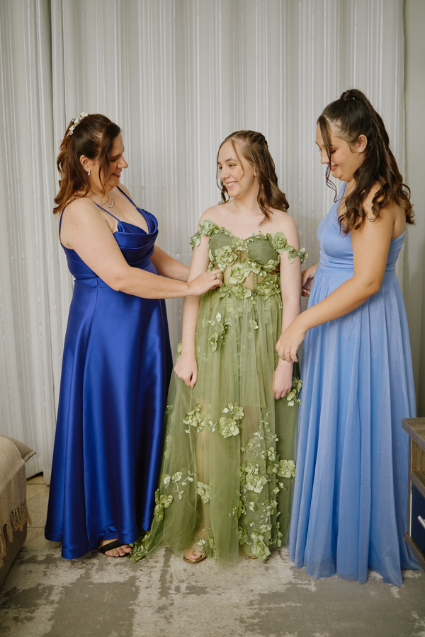 Anna Clara sorri enquanto é auxiliada a vestir seu vestido verde com aplicações de flores. O registro documental captura a cumplicidade e a emoção da debutante em Jacareí, transformando o momento da preparação em uma memória artística e verdadeira.