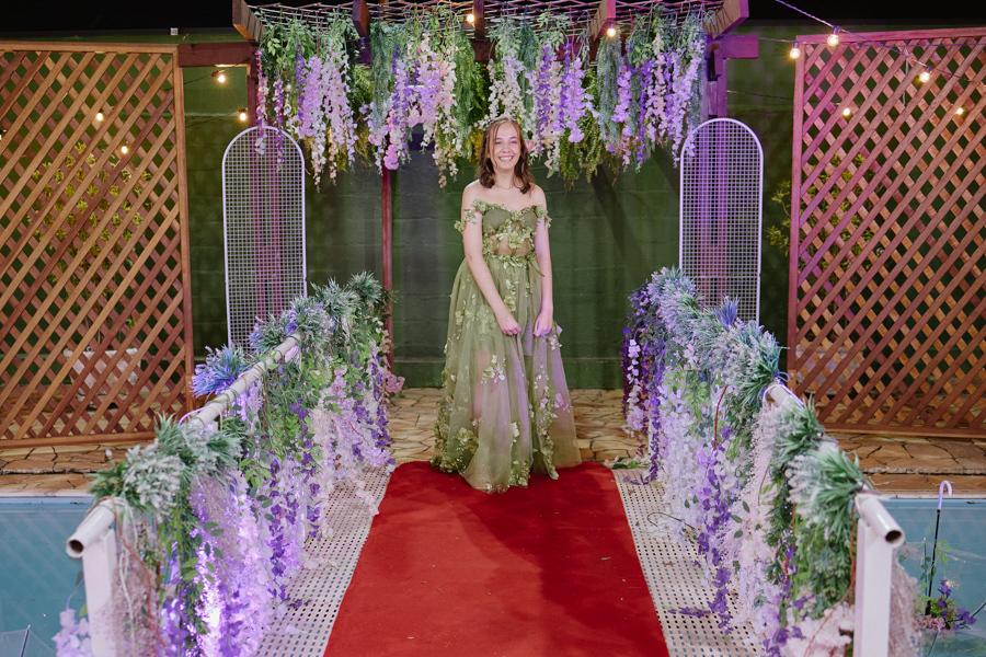Anna Clara caminha radiante pelo tapete vermelho sob um arco de flores lilases. O registro captura a debutante de Jacareí em seu vestido verde artístico, integrando sua essência sensível à decoração encantadora da celebração de seus 15 anos.