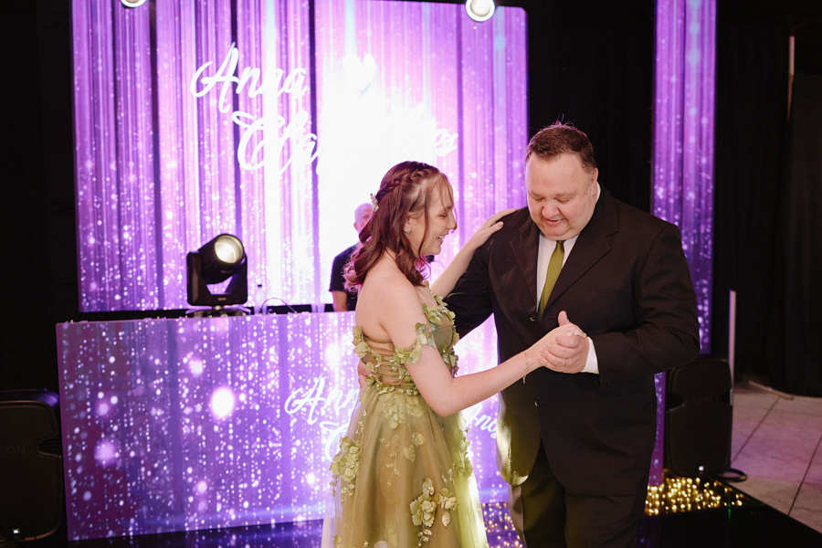 Anna Clara sorri emocionada enquanto dança com seu pai na pista iluminada. A fotografia documental em Jacareí registra a espontaneidade e o afeto real da debutante, emoldurados pelo brilho lilás de uma celebração que é pura expressão de arte e amor.