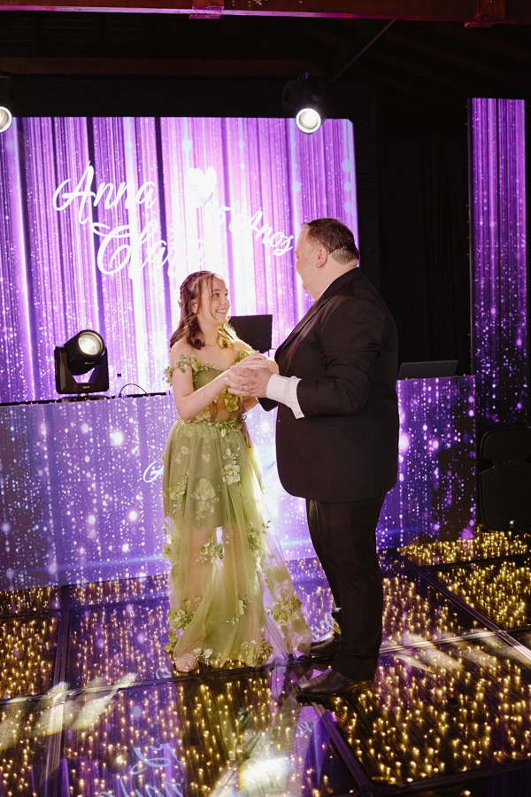 Anna Clara sorri radiante durante a valsa com seu pai na pista de dança iluminada. A fotografia documental em Jacareí captura a alegria genuína e a essência artística da debutante, emoldurada por luzes e painéis em tons de roxo.