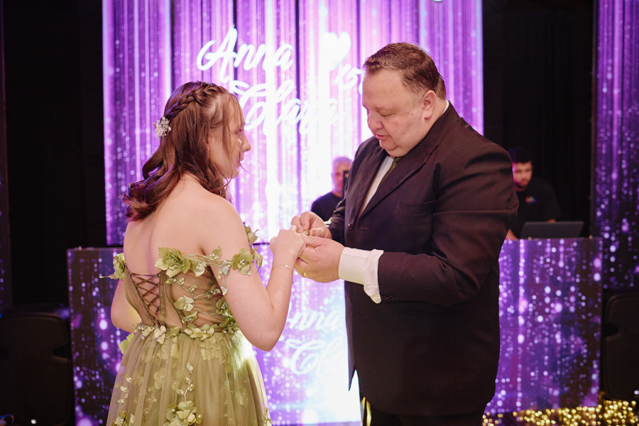 Anna Clara recebe o anel de seu pai durante a cerimônia de 15 anos. A fotografia documental em Jacareí capta a entrega e a emoção deste gesto tradicional, celebrando a identidade sensível da debutante em um cenário artístico iluminado.