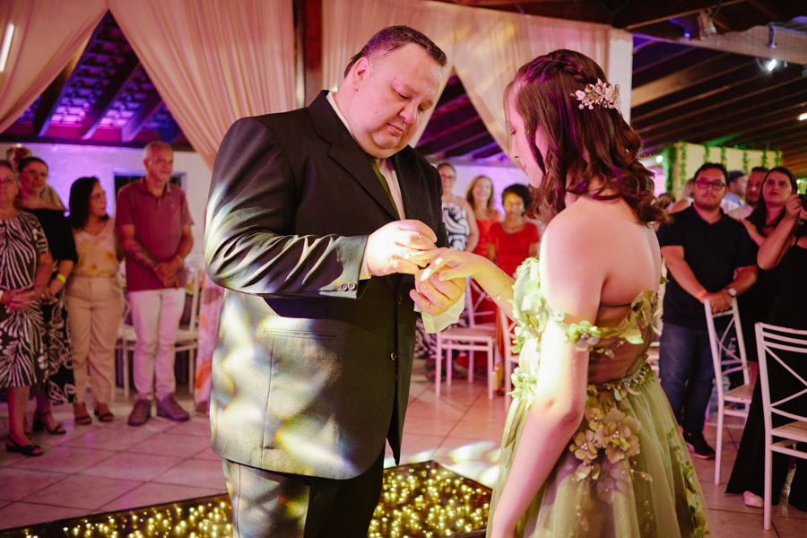 Pai coloca o anel no dedo de Anna Clara durante a cerimônia de 15 anos em Jacareí. A fotografia documental registra a emoção solene e a identidade sensível da debutante, destacando o detalhe artístico de seu vestido verde e a iluminação acolhedora.