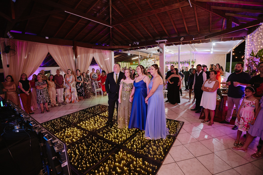 Anna Clara e sua família assistem a uma homenagem na pista de dança iluminada. A fotografia documental em Jacareí registra a união e a emoção dos 15 anos, capturando a atmosfera artística e o carinho dos convidados sob a luz quente da celebração.