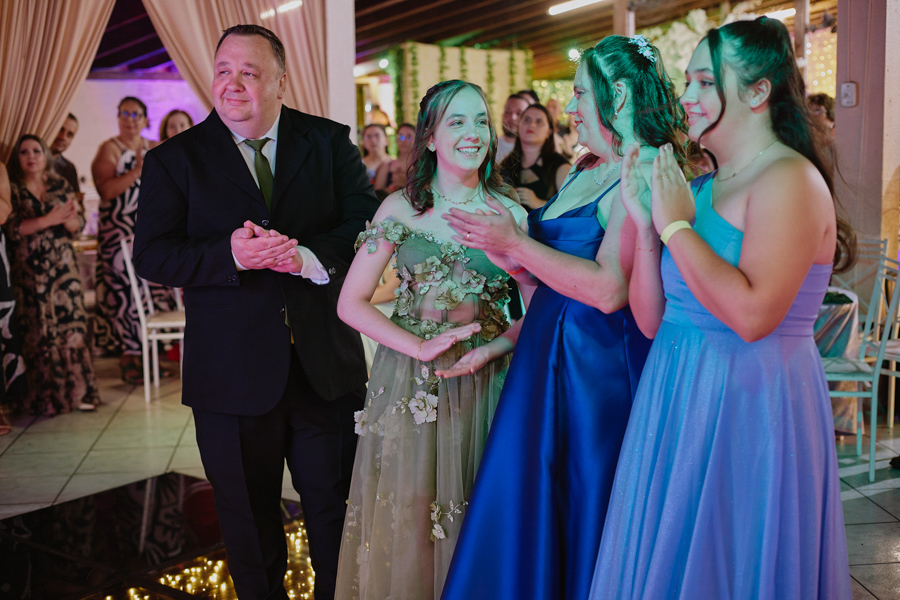 Anna Clara e sua família aplaudem sorridentes durante a celebração de seus 15 anos. A fotografia documental em Jacareí captura a alegria genuína e a cumplicidade desse momento, refletindo a essência sensível e artística da debutante e seus pais.