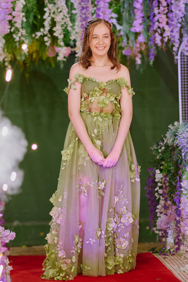Retrato de Anna Clara sorridente em seu vestido verde artístico com aplicações florais. A fotografia documental em Jacareí destaca a sensibilidade e a alegria da debutante, emoldurada por uma decoração floral que reflete sua identidade criativa.