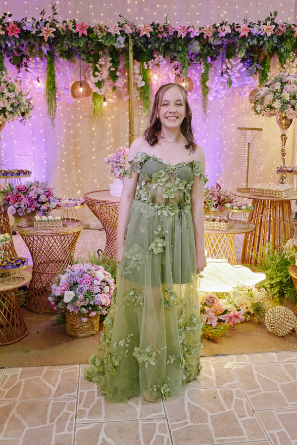 Anna Clara sorri diante da mesa de doces decorada com flores e luzes. O registro documental em Jacareí destaca a debutante em seu vestido verde artístico, capturando a harmonia entre sua essência sensível e a atmosfera encantadora de seus 15 anos.