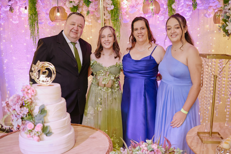 Anna Clara posa sorridente com sua família ao lado do bolo de 15 anos. A fotografia documental em Jacareí registra a união e o afeto real, destacando o cenário artístico e iluminado que reflete a identidade sensível da debutante e sua celebração.