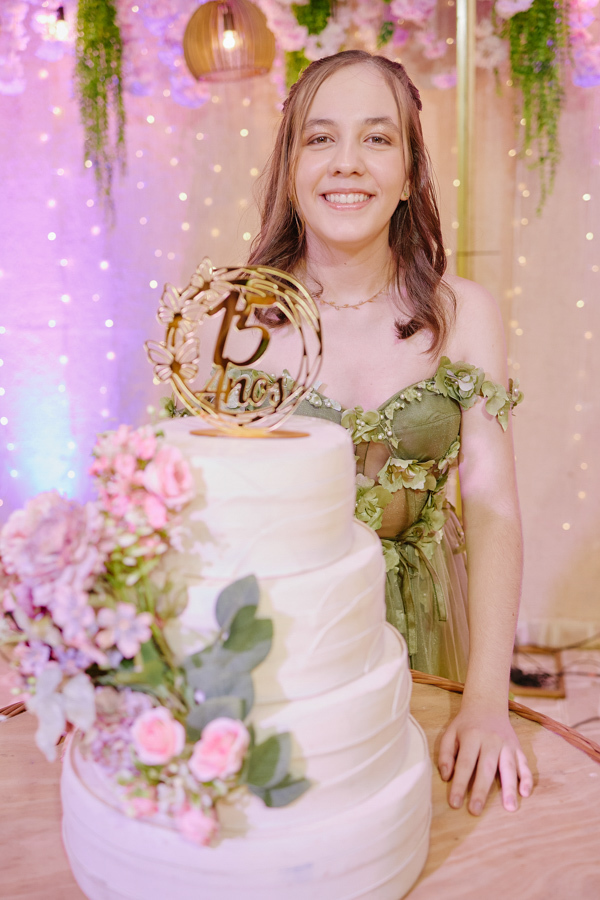 Anna Clara sorri radiante posicionada atrás de seu bolo de 15 anos. A fotografia documental em Jacareí destaca a alegria e a sensibilidade da debutante, capturando sua essência artística em um cenário iluminado e acolhedor de celebração.