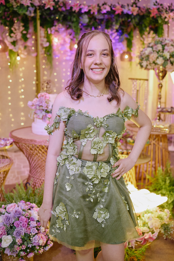 Anna Clara sorri com confiança em seu vestido verde artístico, agora em versão curta para a recepção. A fotografia documental em Jacareí destaca sua essência vibrante e sensível, emoldurada pela decoração floral e iluminada de seus 15 anos.