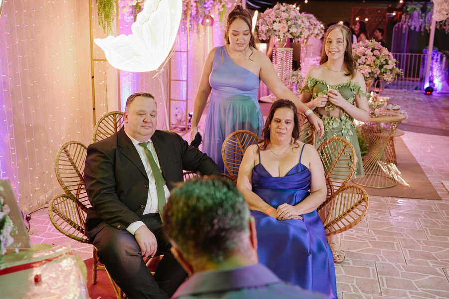 Anna Clara e sua irmã observam atentamente enquanto seus pais posam para um caricaturista. O registro documental em Jacareí captura a integração da família à proposta artística da festa de 15 anos, em um cenário acolhedor e cheio de significado.