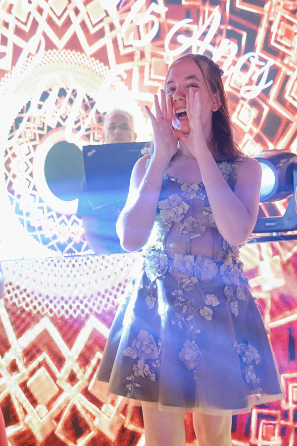 Anna Clara grita de alegria com as mãos no rosto em frente ao telão de LED. O registro documental em Jacareí captura a euforia e a energia da debutante na pista de dança, destacando sua essência vibrante e o clima festivo de seus 15 anos.