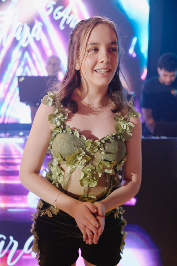 Anna Clara sorri com brilho no olhar durante a recepção de seus 15 anos. A fotografia documental em Jacareí destaca a debutante em seu traje artístico de festa, capturando sua alegria e espontaneidade em meio à energia vibrante da pista de dança.