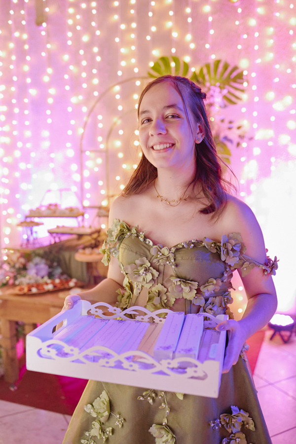 Anna Clara sorri segurando uma bandeja com lembranças para seus convidados. O registro documental em Jacareí destaca a delicadeza e a gratidão da debutante, emoldurada por um fundo de luzes brilhantes que reforça a magia de seus 15 anos.