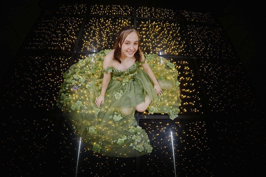 Vista aérea de Anna Clara sentada sobre a pista de LED iluminada, com seu vestido verde artístico espalhado ao redor. O registro documental em Jacareí captura a magia e a beleza da debutante em um momento poético e brilhante de seus 15 anos.