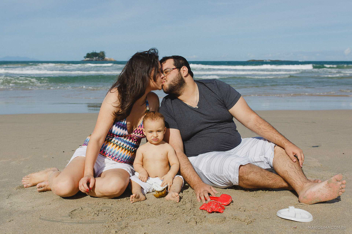 Beijo dos pais sentados na areia junto com o filho em praia do Guarujá