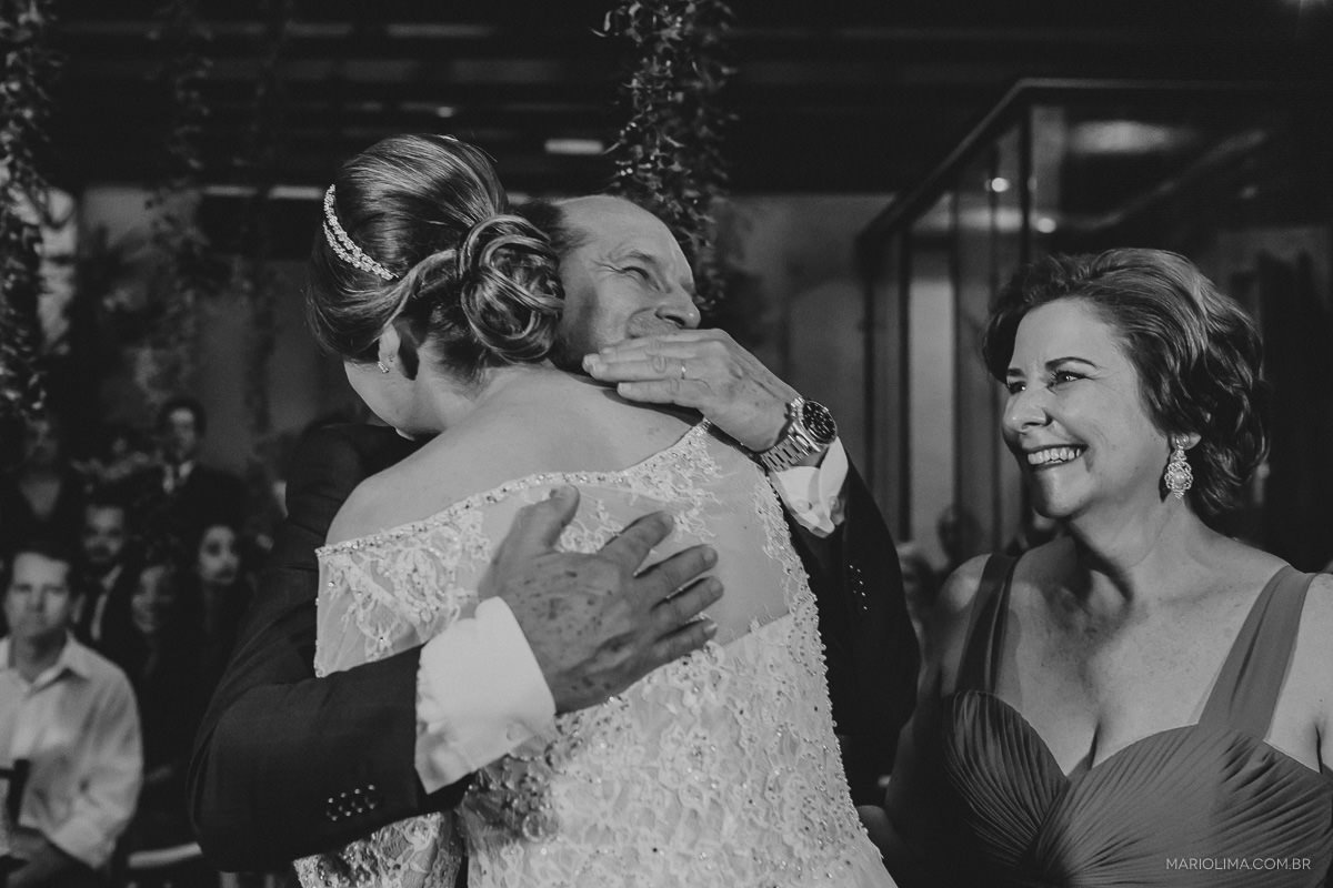 Mãe da noiva sorrindo ao vê-la abraçando o pai em cerimônia de casamento no Espaço Lilló