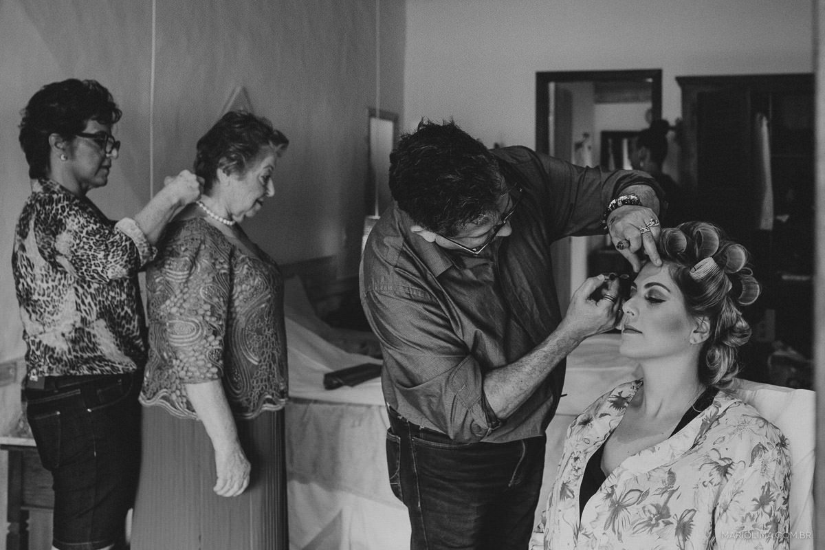 Fotografia de noiva sendo maquiada por Henrique Melo da Agência First e mãe ajudando a avó com o vestido