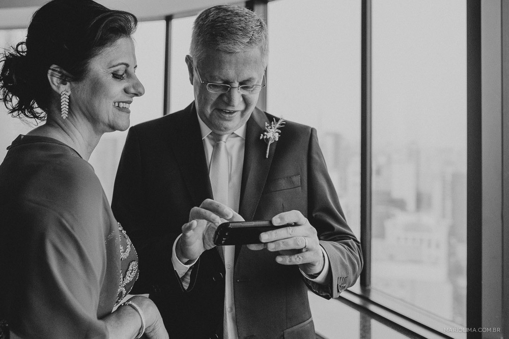 Fotografia do pai da noiva mexendo no celular e mãe da noiva olhando