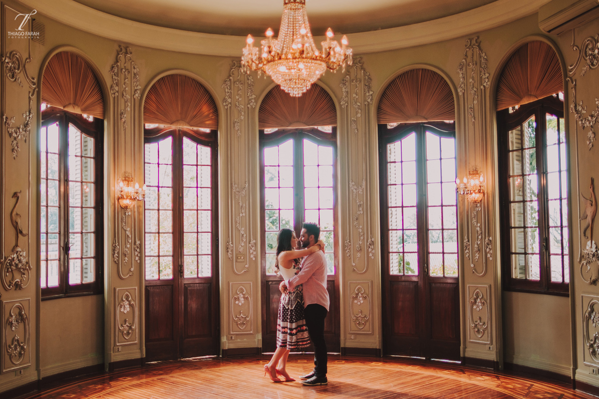 casal juntos abarçados, no meio da sala, com várias janelas, lustre e arquitetura vitoriana.