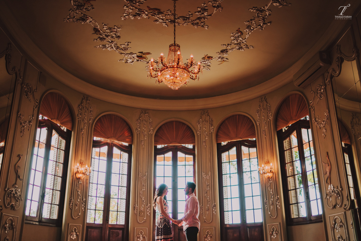 casal de mãos dadas no meio da sala, com várias janelas, lustre e arquitetura vitoriana.