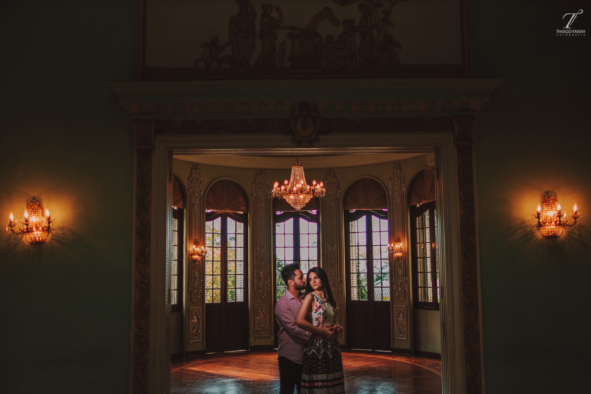 casal juntos abarçados, no meio da sala, com várias janelas, lustre e arquitetura vitoriana.