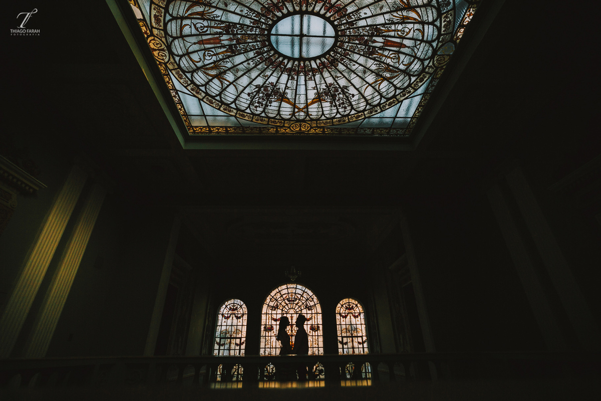casal juntos abarçados, no meio da sala, com várias janelas, e arquitetura vitoriana. foto contra luz.