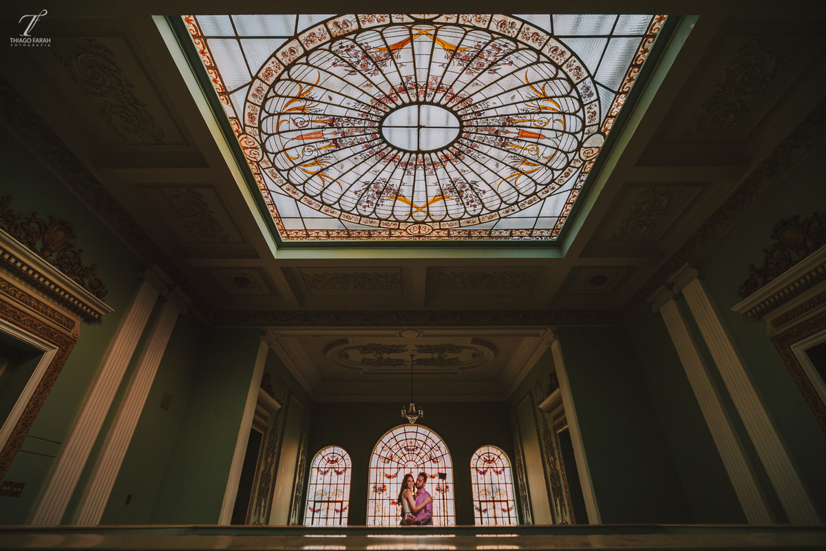 casal juntos abarçados, no meio da sala, com várias janelas, e arquitetura vitoriana.