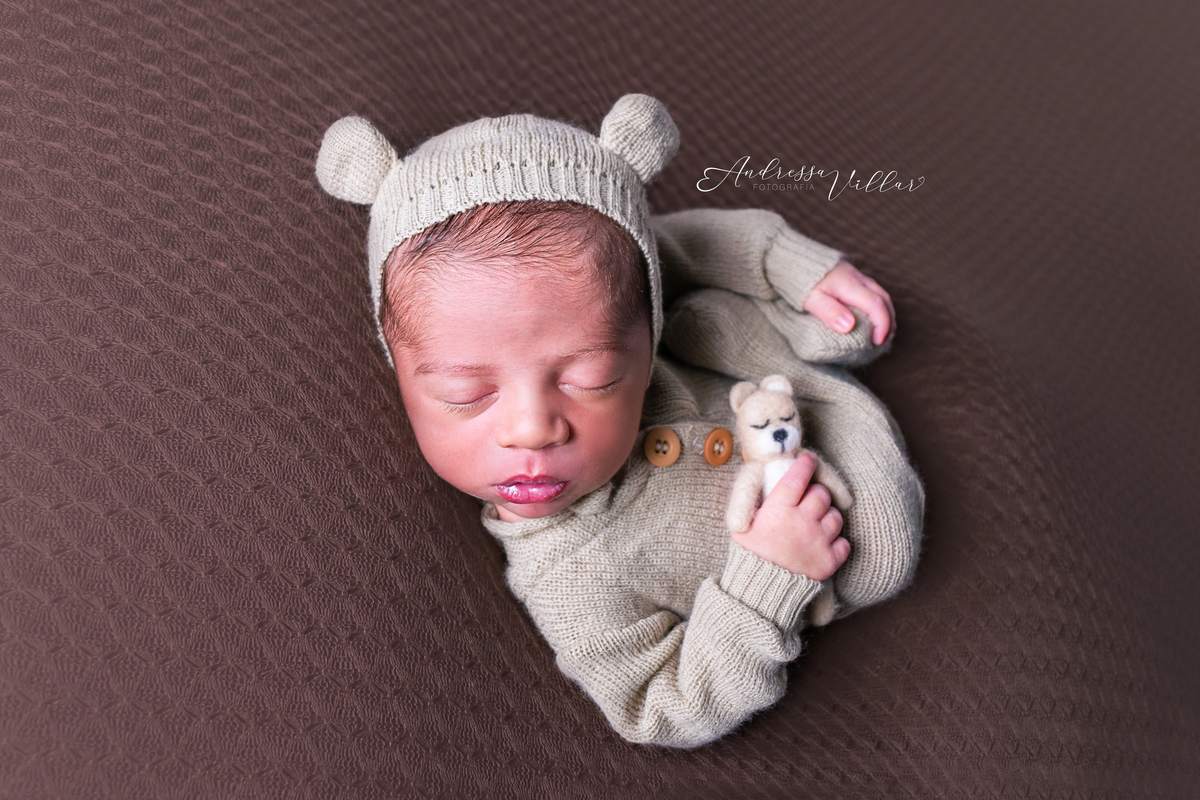 Recém Nascido deitado de barriga para cima com touca de ursinho da cor do macacão, segurando um ursinho