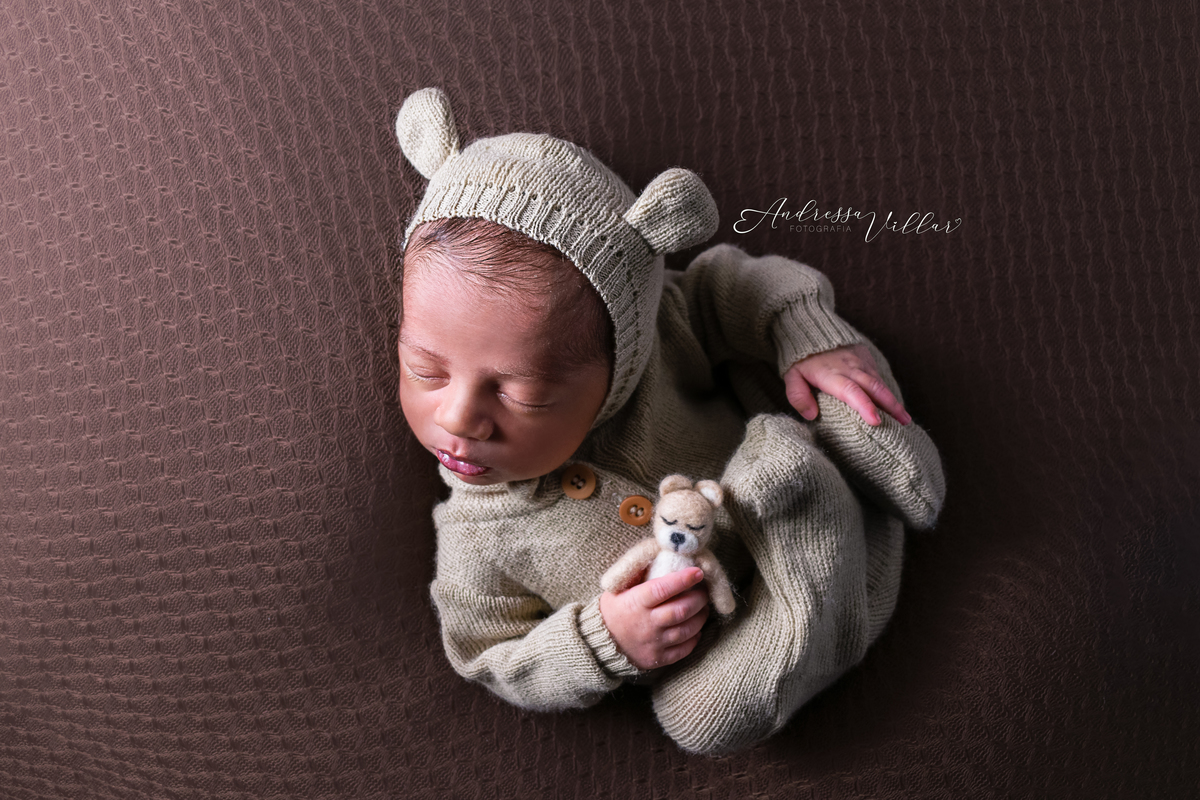 Recém Nascido deitado de barriga para cima com touca de ursinho da cor do macacão, segurando um ursinho