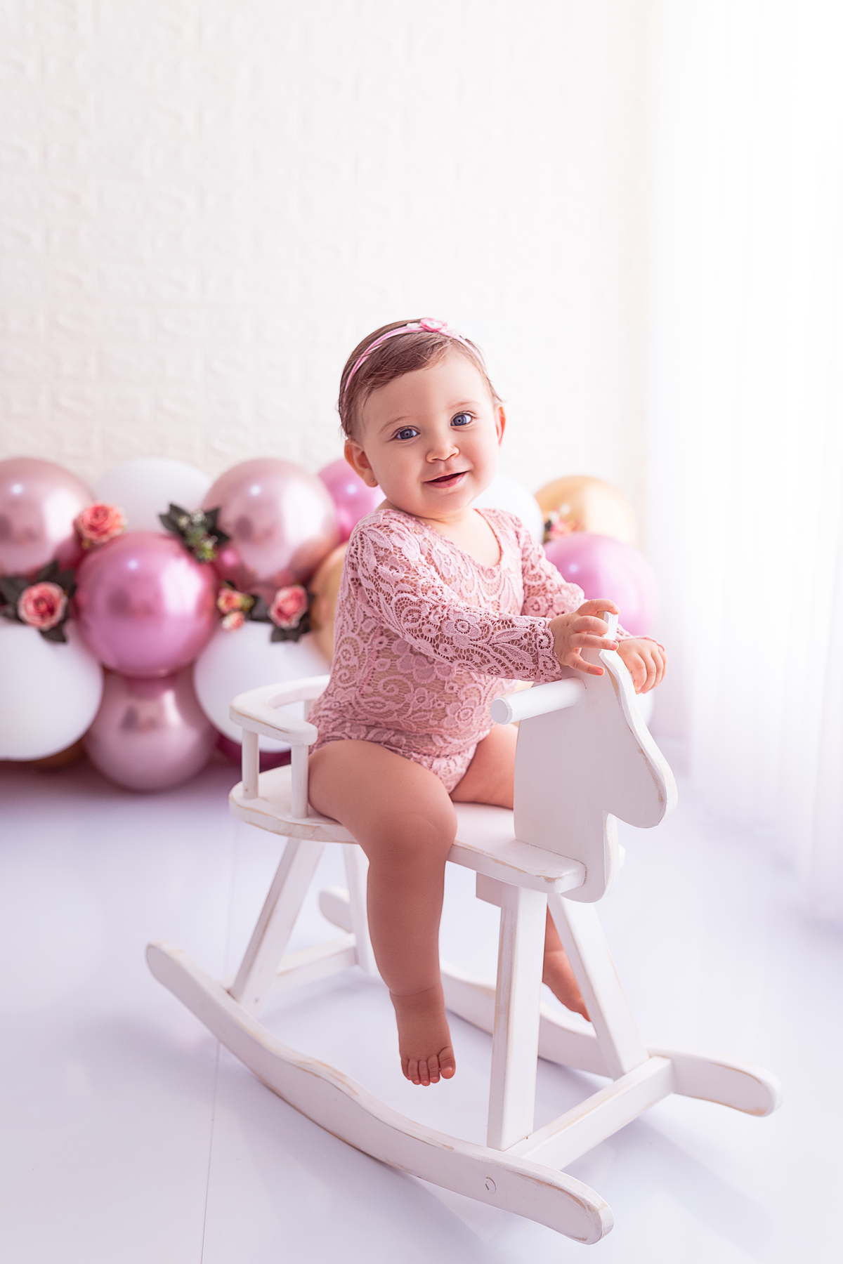 Giovanna sentada em um cavalinho branco de madeira vestindo body rendado rosa com balões rosa e dourado e flores ao fundo