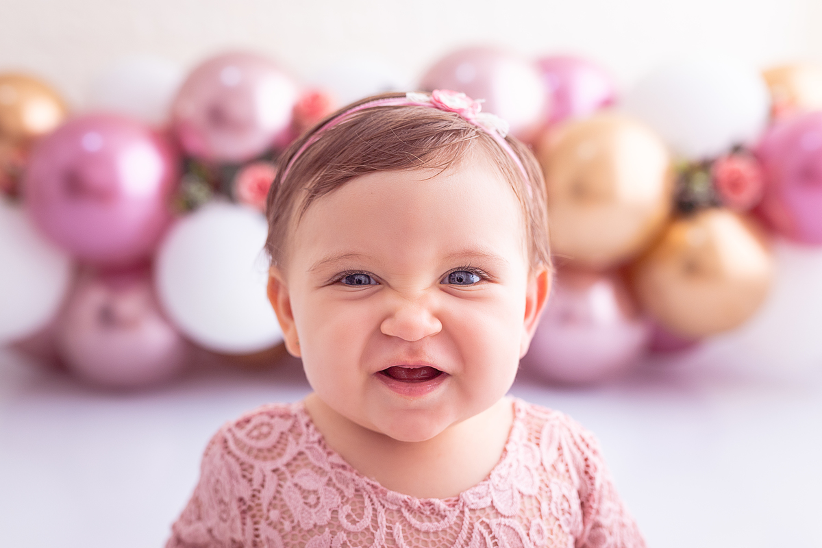 Giovanna  sorrindo e vestindo body rendado rosa com balões rosa e dourado e flores ao fundo
