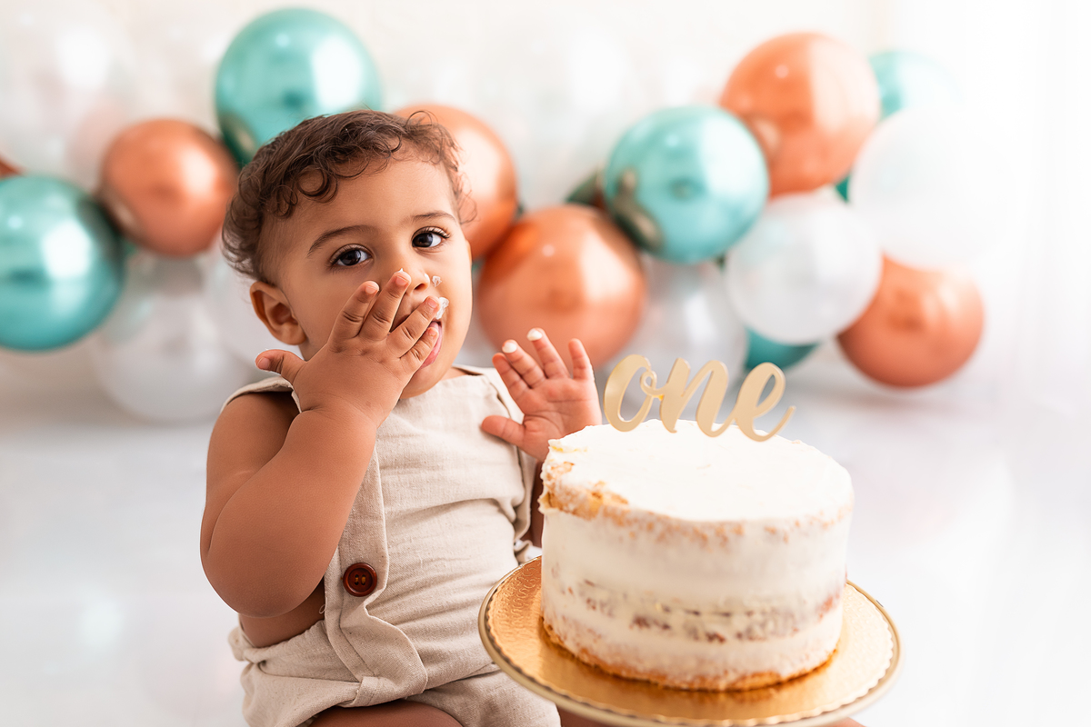 bebê lambendo a mão suja com bolo sem açúcar e balões ao fundo