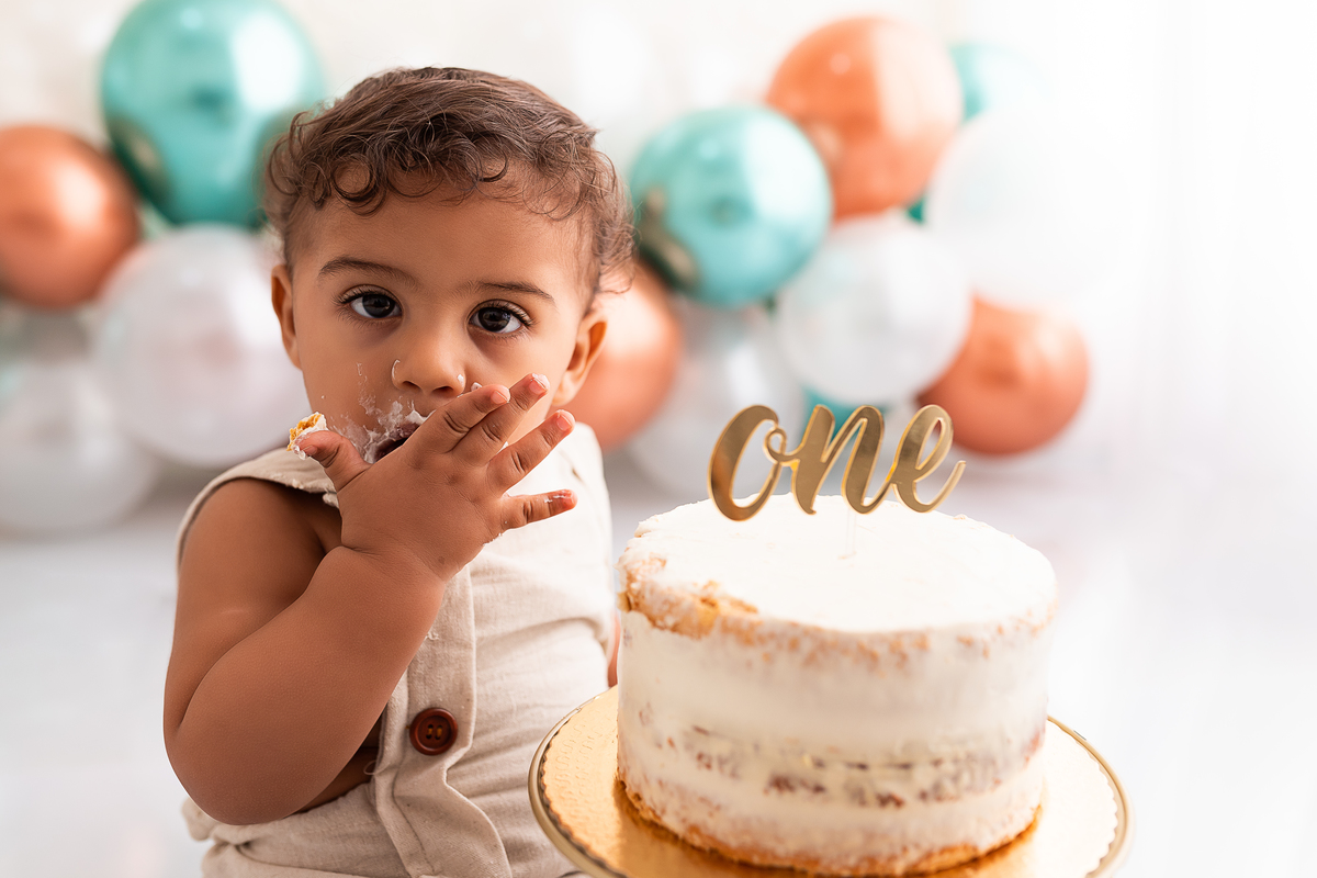 bebê lambendo a mão suja com bolo sem açúcar e balões ao fundo