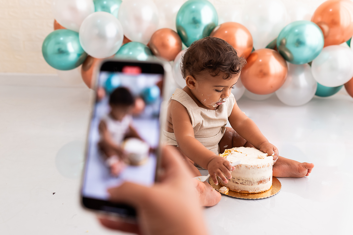 Mamãe usando o celular para fotografar o bebê se lambuzando com o bolo 