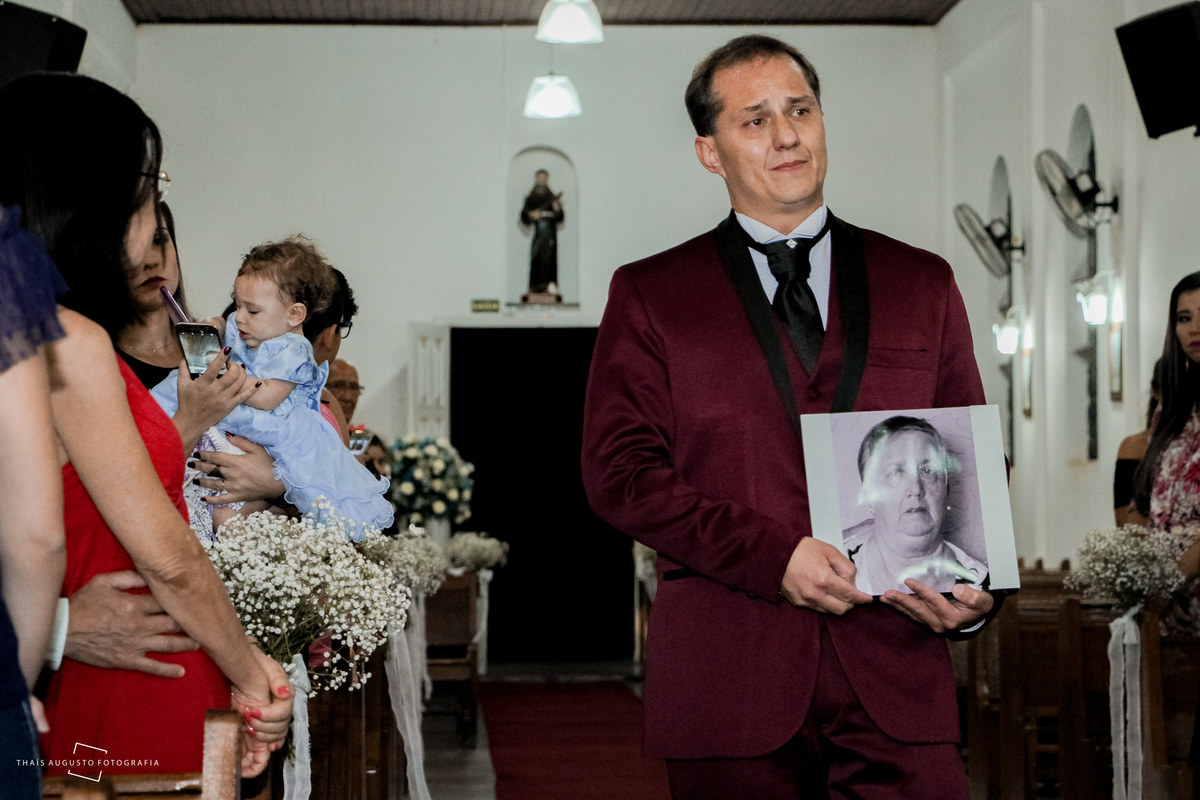 noivo faz homenagem a mãe falecida na entrada do casamento, terno marsala, casamento realizado na cidade de agudos, igreja santo antônio casamento católico fotógrafa de casamento thais augusto fotografia registra momento especial.