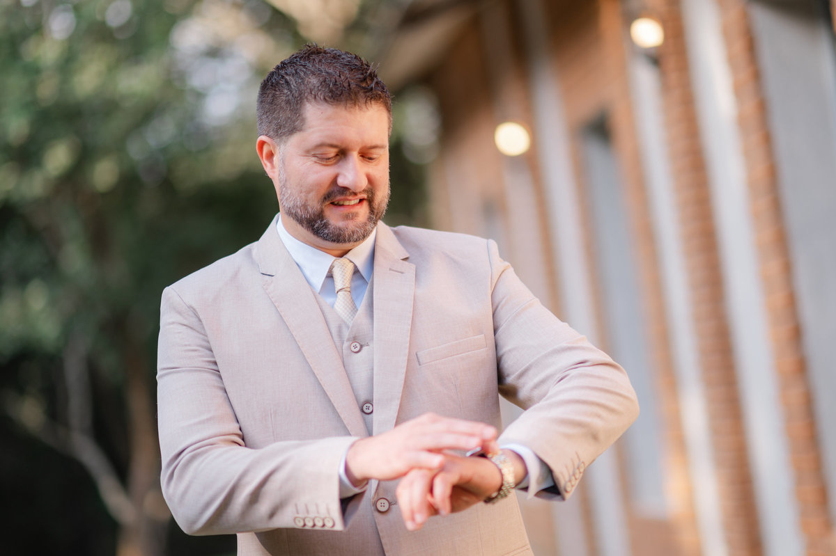 Juliano ajusta o relógio durante os preparativos para seu casamento, em um retrato que destaca a elegância do traje.