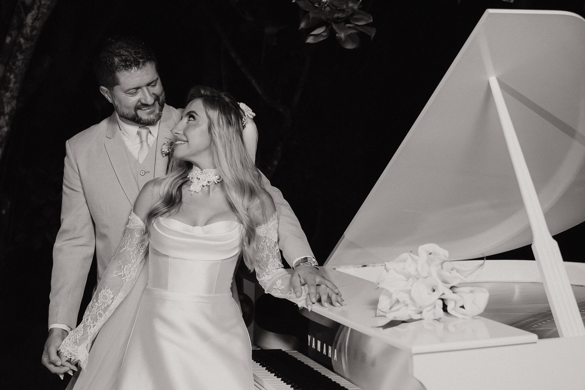 Retrato de Ana Rute e Juliano junto ao piano branco, em uma composição elegante durante a recepção do casamento.