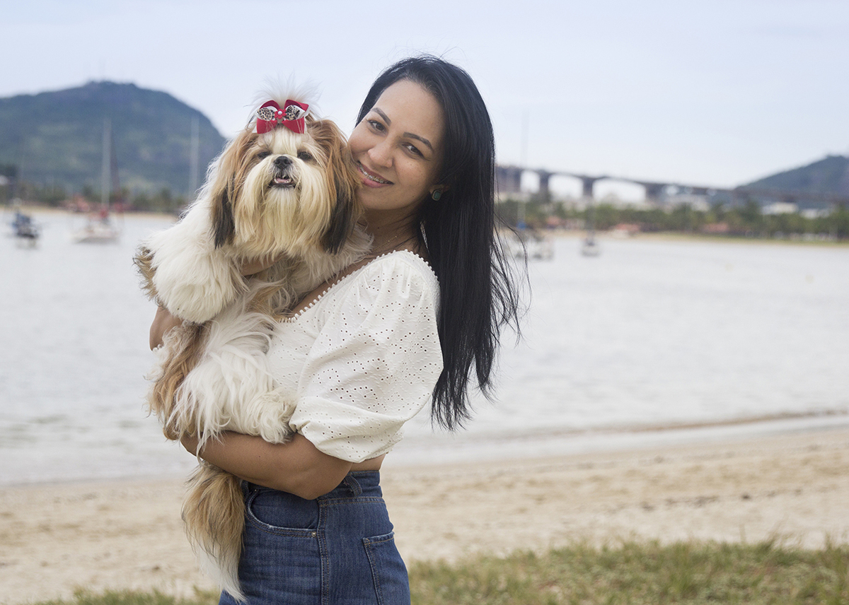 Shih-tzu no colo da tutora, na praia, com terceira ponte ao fundo
