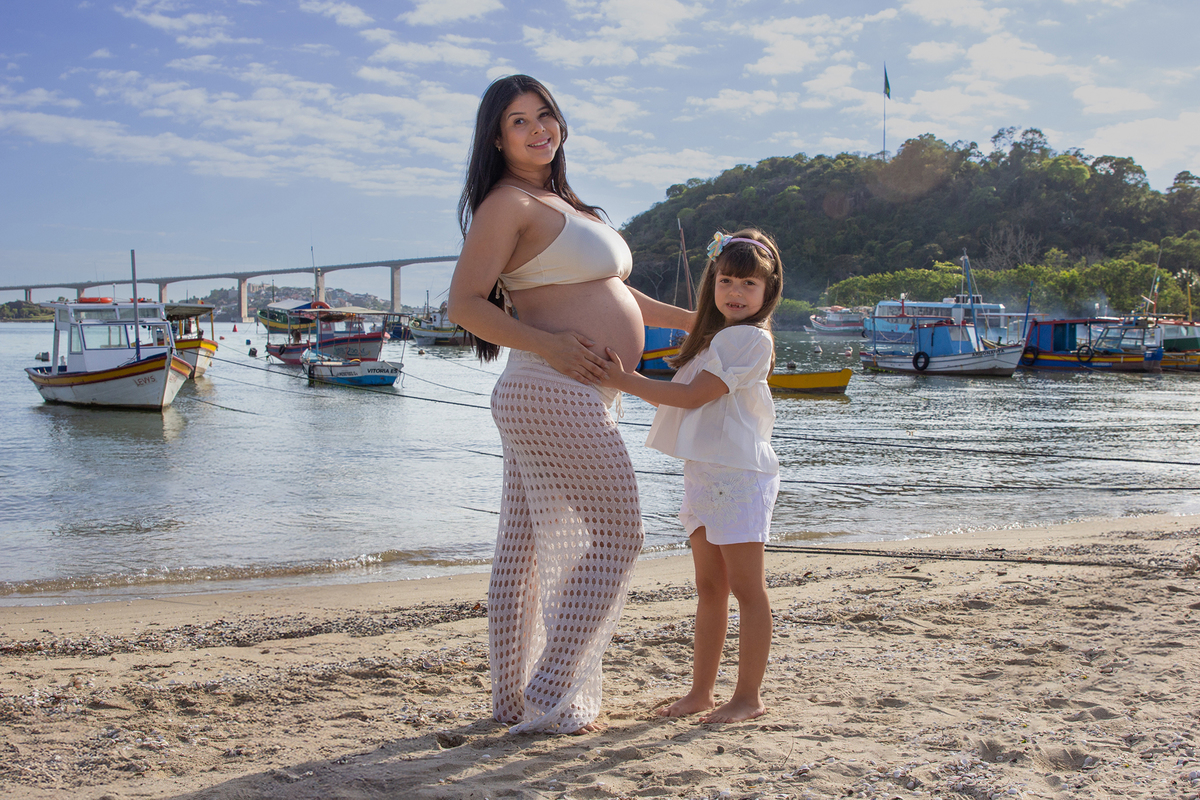mãe e filha, olhando para barriga, na praia, com mar e terceira ponte ao fundo 